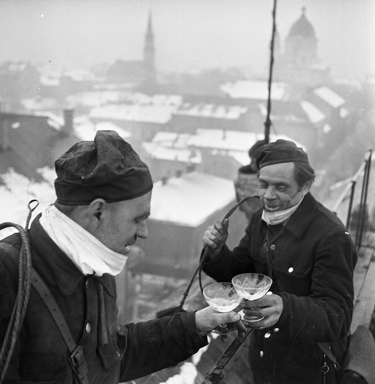 Two chimney sweeps in caps and scarves toast with glasses on a snowy rooftop above a town in 1970