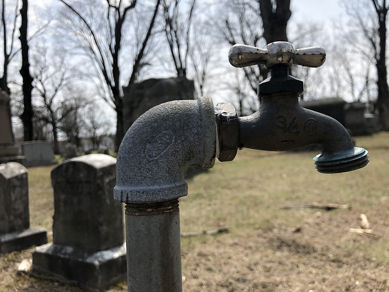 An old outdoor hose spigot with a cross handle on a galvanized standpipe, bare winter trees behind it