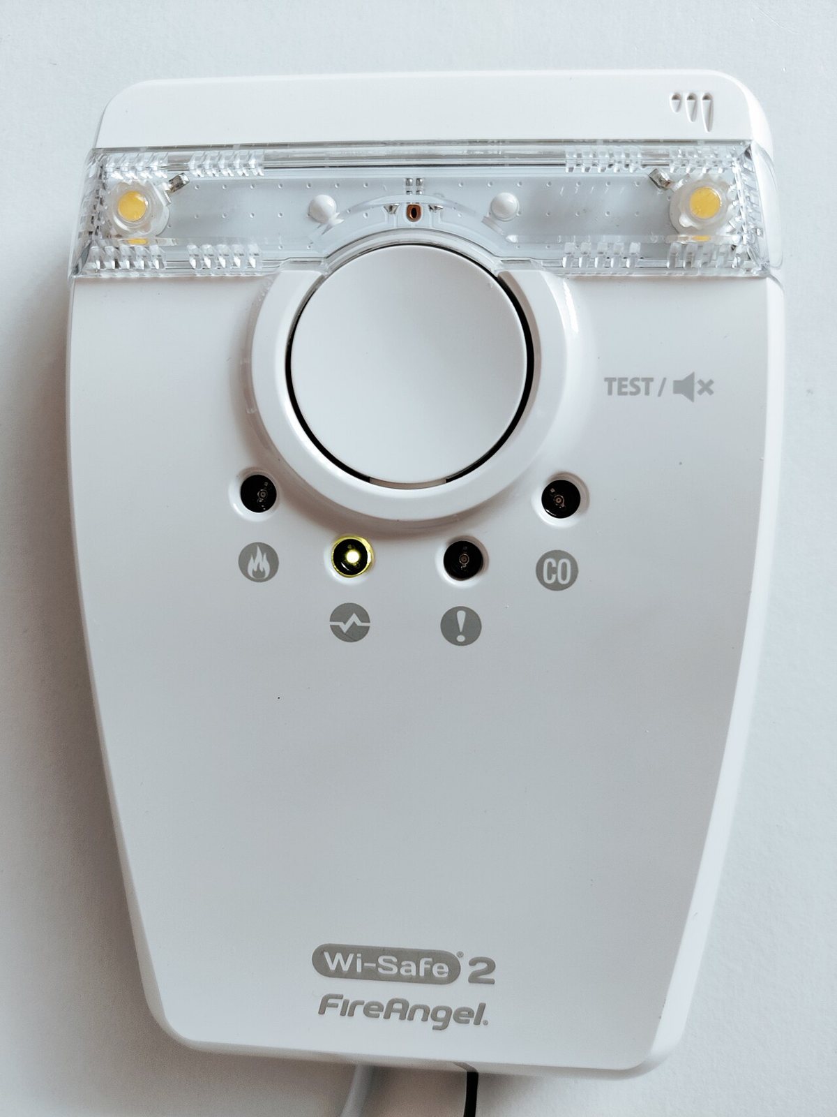 A white wall-mounted alarm control unit with a large round test button and small indicator lights marked for fire and CO