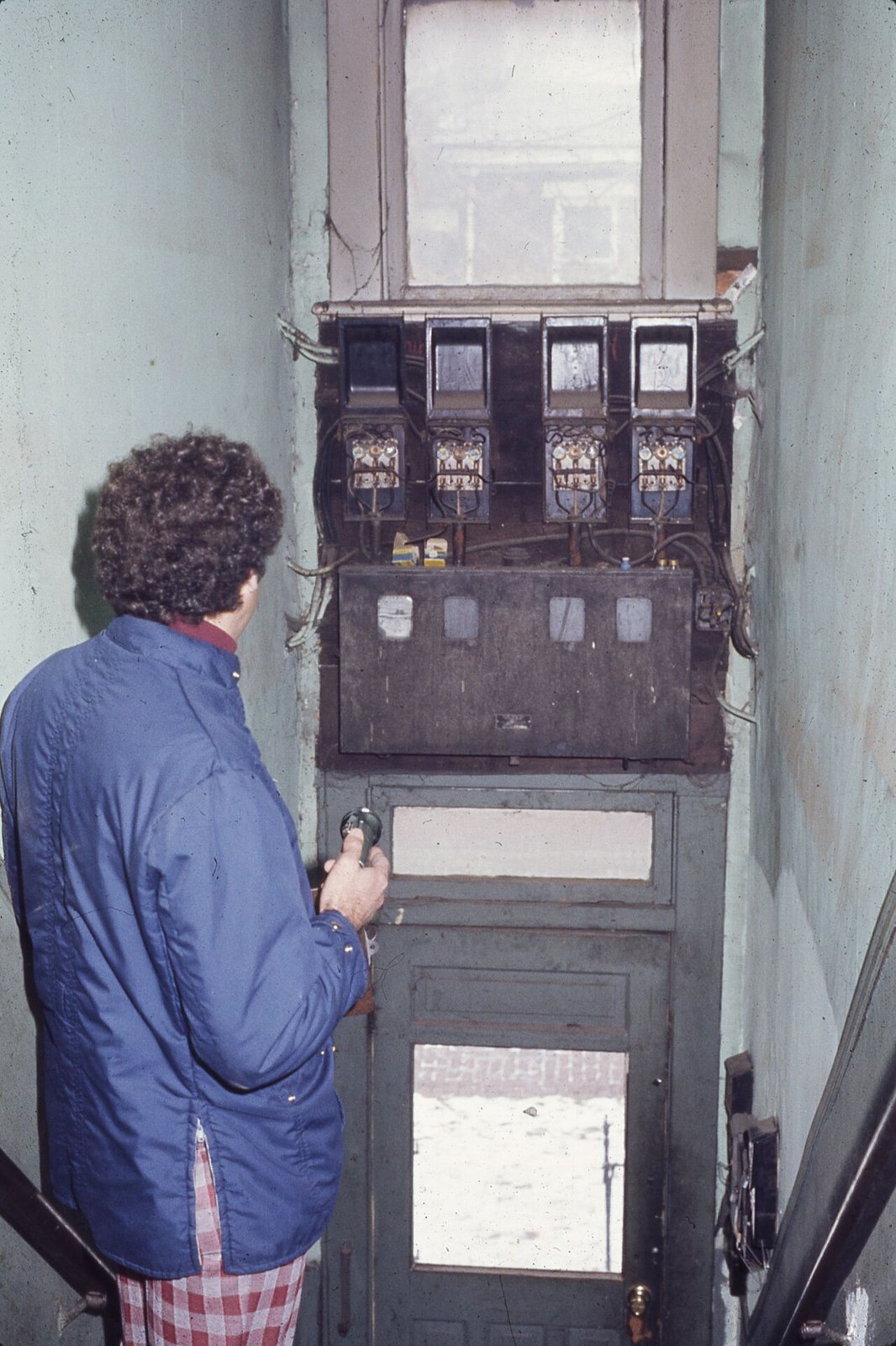 An inspector in a blue jacket holds a flashlight while looking at an old wall of electric meters and fuse boxes above a doorway